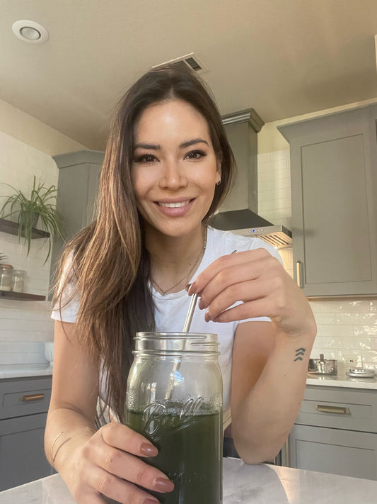 Woman smiling and stirring a green drink in a mason jar in a bright kitchen