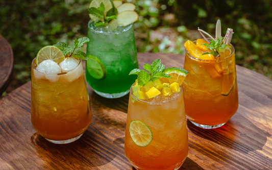 Four colorful non-alcoholic cocktails arranged on a wooden table outdoors, garnished with lime, mint, mango and lychee