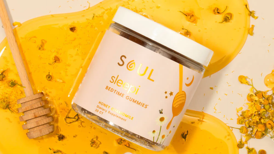 Angled jar of Soul Sleepi bedtime gummies with pink label and white lid on spilled honey, a wooden honey dipper, and chamomile flowers