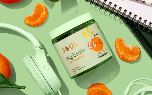 Jar of SOUL Big Brain Focus Gummies (tangerine) next to headphones, tangerine slices, and a notebook on a green background
