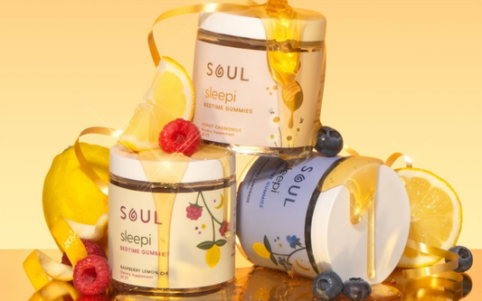 Three-quarter view of three Soul Sleepi Bedtime Gummies jars with honey drizzle, lemon slices, raspberries, and blueberries on a warm yellow studio background