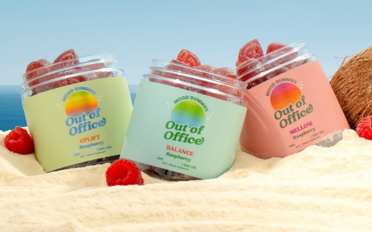 Angled view of three Out of Office mood gummy jars (Uplift, Balance, Mellow) with raspberry gummies on sand and ocean background