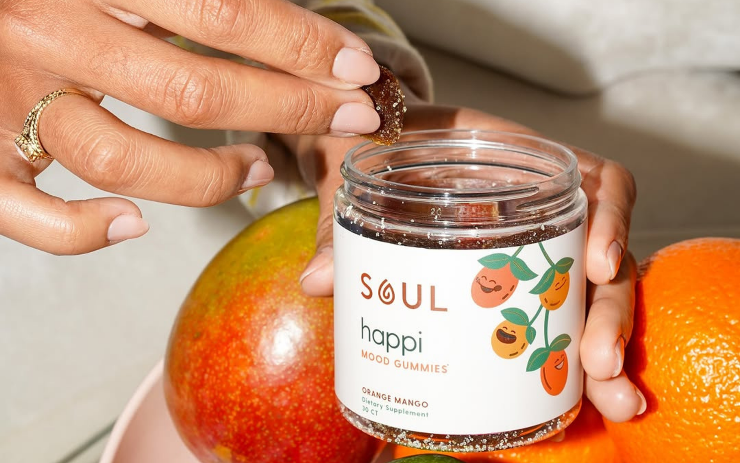 Hand picking a Soul happi Orange Mango mood gummy from an open jar beside mango and orange