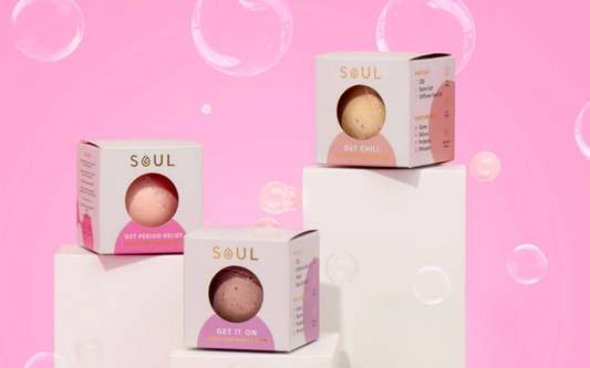 Three SOUL CBD bath bomb boxes with bath bombs visible in windowed boxes on pedestals against a pink background with bubbles