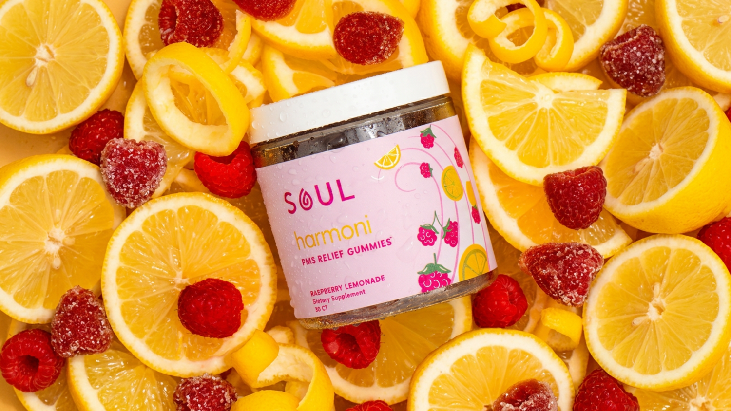 Angled jar of SOUL Harmoni PMS Relief Gummies showcasing pink label, raspberry lemonade flavor, condensation, with lemon slices and raspberries background
