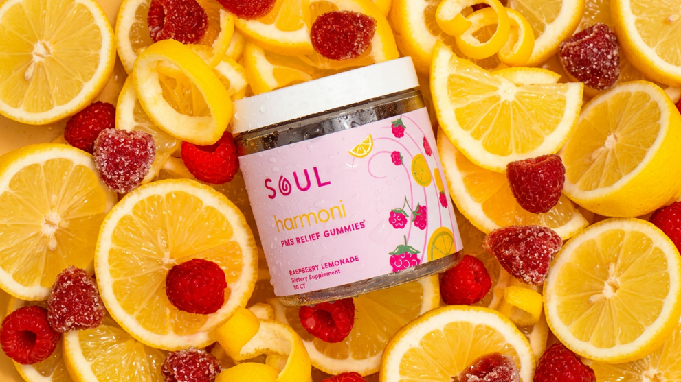 Angled jar of SOUL Harmoni PMS Relief Gummies showcasing pink label, raspberry lemonade flavor, condensation, with lemon slices and raspberries background