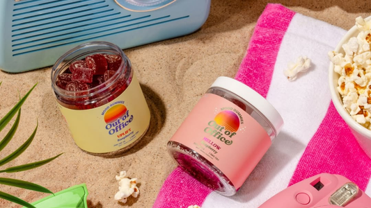 Angled view of two Out of Office mood gummies jars showcasing red gummies and labeled Uplift raspberry and Mellow strawberry on sand and a pink-striped towel