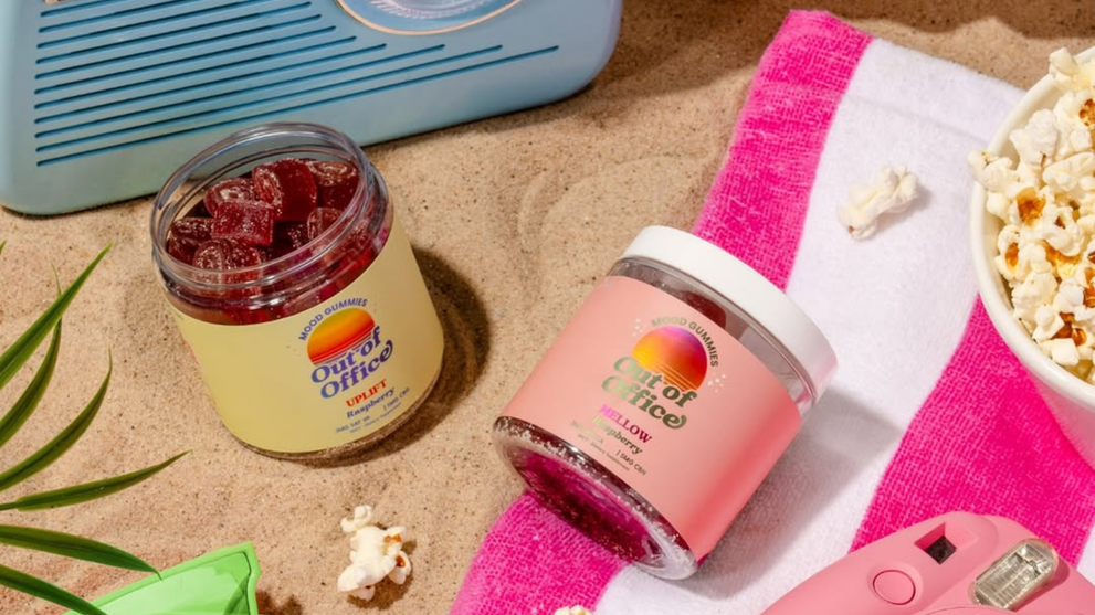 Angled view of two Out of Office mood gummies jars showcasing red gummies and labeled Uplift raspberry and Mellow strawberry on sand and a pink-striped towel