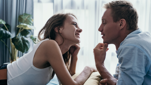 Couple leaning toward each other and smiling on a bed in a softly lit bedroom
