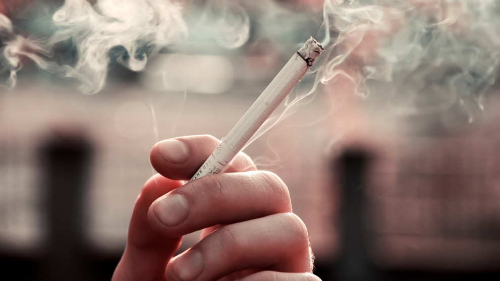 Hand holding a lit cigarette with smoke curling upward against a blurred outdoor background